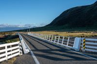 a white road next to a grassy hillside with mountains in the distance, with fence posts and grass along one side