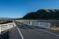 a white road next to a grassy hillside with mountains in the distance, with fence posts and grass along one side