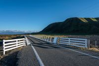 a white road next to a grassy hillside with mountains in the distance, with fence posts and grass along one side