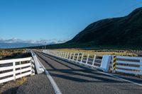 a white road next to a grassy hillside with mountains in the distance, with fence posts and grass along one side