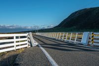 a white road next to a grassy hillside with mountains in the distance, with fence posts and grass along one side