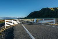 a white road next to a grassy hillside with mountains in the distance, with fence posts and grass along one side