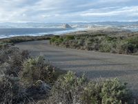 a road going to the beach towards a body of water and mountains, surrounded by scrub