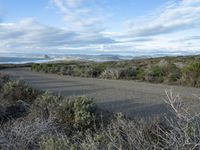 a road going to the beach towards a body of water and mountains, surrounded by scrub