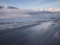 there is some ice on the road next to mountains with snow and clouds covering them