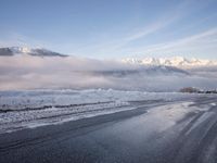 there is some ice on the road next to mountains with snow and clouds covering them