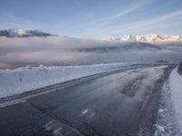 there is some ice on the road next to mountains with snow and clouds covering them
