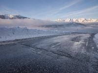 there is some ice on the road next to mountains with snow and clouds covering them