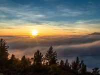 clouds and trees surround an area with sunsets and mountains in the distance that is covered by low lying clouds