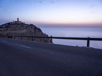 a lighthouse is sitting on top of the hill above the ocean at dusk with a car driving along it