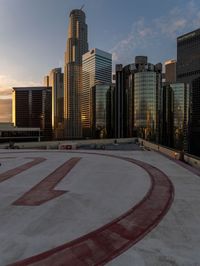 a man walking across an empty parking lot towards a skyline at sunset with high rise buildings in the background