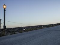 an empty street under a light post near the water and telephone wires, in the distance, is a lighthouse on a stone - built wall