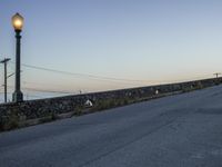 an empty street under a light post near the water and telephone wires, in the distance, is a lighthouse on a stone - built wall