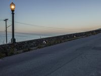 an empty street under a light post near the water and telephone wires, in the distance, is a lighthouse on a stone - built wall