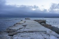 a very wide rocky pier with benches at the end of it on the water surface