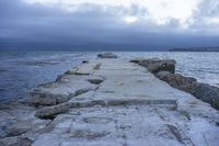 a very wide rocky pier with benches at the end of it on the water surface