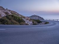 a curved road near a lighthouse, overlooking the ocean at sunset from below it is visible