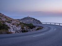 a curved road near a lighthouse, overlooking the ocean at sunset from below it is visible