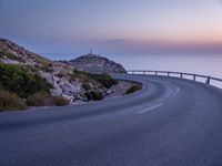 a curved road near a lighthouse, overlooking the ocean at sunset from below it is visible