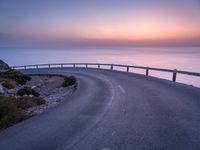 a curved road near a lighthouse, overlooking the ocean at sunset from below it is visible