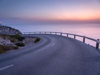 a curved road near a lighthouse, overlooking the ocean at sunset from below it is visible