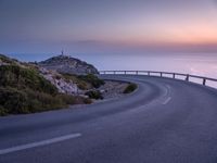 a curved road near a lighthouse, overlooking the ocean at sunset from below it is visible