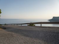 a road in front of a cliff overlooking the water and coastline with a bench next to it