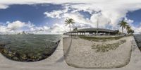 the same fish eye view is showing a pier on an ocean shore and palm trees on an island in the background