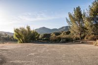 a wide empty parking lot next to trees in front of a mountain range and blue sky