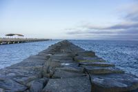 the stone wall leading out into the ocean with pier in background and overcast sky