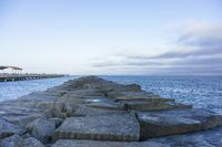 the stone wall leading out into the ocean with pier in background and overcast sky