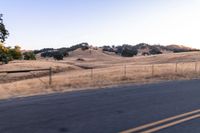 a motorcycle traveling along a rural road with hills in the background and a fence separating the middle