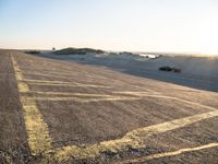 an empty road with lines painted in the sand near a beach and ocean with cars going to a left