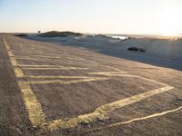 an empty road with lines painted in the sand near a beach and ocean with cars going to a left