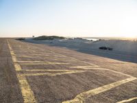 an empty road with lines painted in the sand near a beach and ocean with cars going to a left