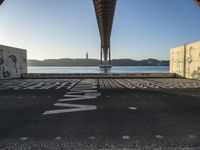 graffiti in street next to a bridge with words on it by the ocean and a road