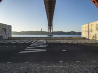 graffiti in street next to a bridge with words on it by the ocean and a road