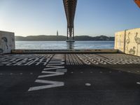 graffiti in street next to a bridge with words on it by the ocean and a road