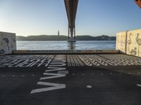 graffiti in street next to a bridge with words on it by the ocean and a road