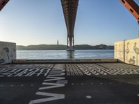 graffiti in street next to a bridge with words on it by the ocean and a road