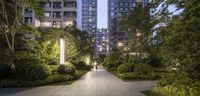 a pathway that has some plants and trees in it and a building with lights on each end