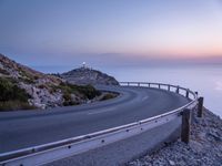 a winding mountain road leading off into the ocean and sunset over it is lit up