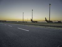 two airplanes parked in a parking lot at sunset behind a runway with poles and light posts