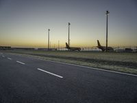 two airplanes parked in a parking lot at sunset behind a runway with poles and light posts