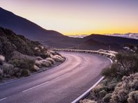 an empty winding road on a mountain with the sun set above the mountains in the background