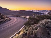 an empty winding road on a mountain with the sun set above the mountains in the background