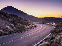 an empty winding road on a mountain with the sun set above the mountains in the background