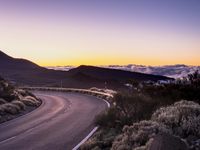 an empty winding road on a mountain with the sun set above the mountains in the background