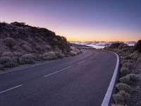 an empty winding road on a mountain with the sun set above the mountains in the background