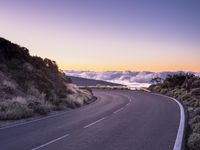 an empty winding road on a mountain with the sun set above the mountains in the background